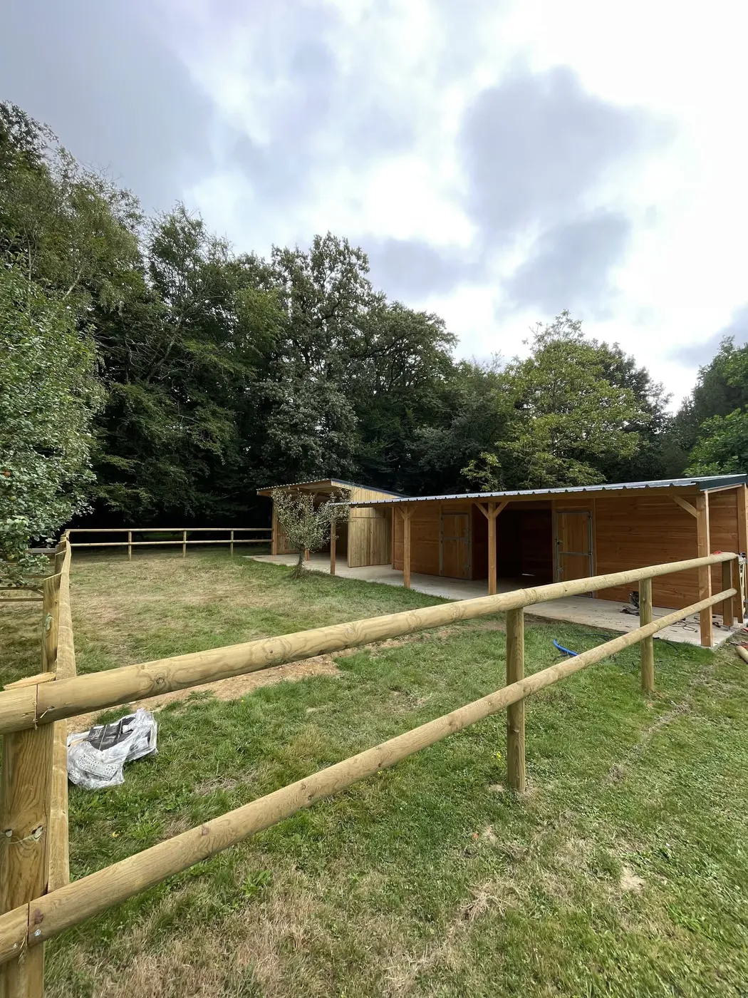 Clôture bois à lices devant un abri et des boxes à chevaux